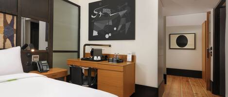 A hotel room with a bed, a desk with a telephone, a coffee machine, and a painting on the wall.