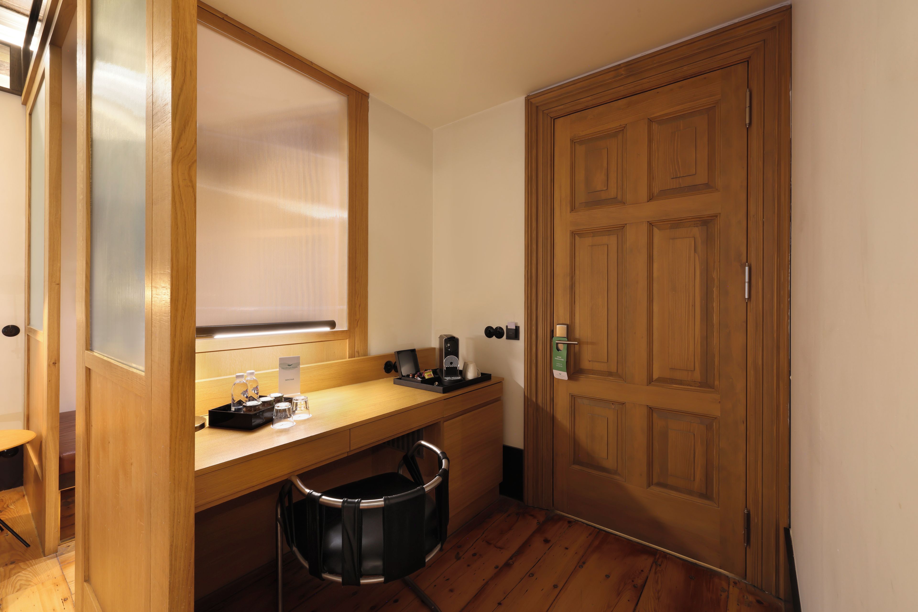 A hotel room with a wooden desk, a coffee maker, a trash bin, and a door with a keycard lock.