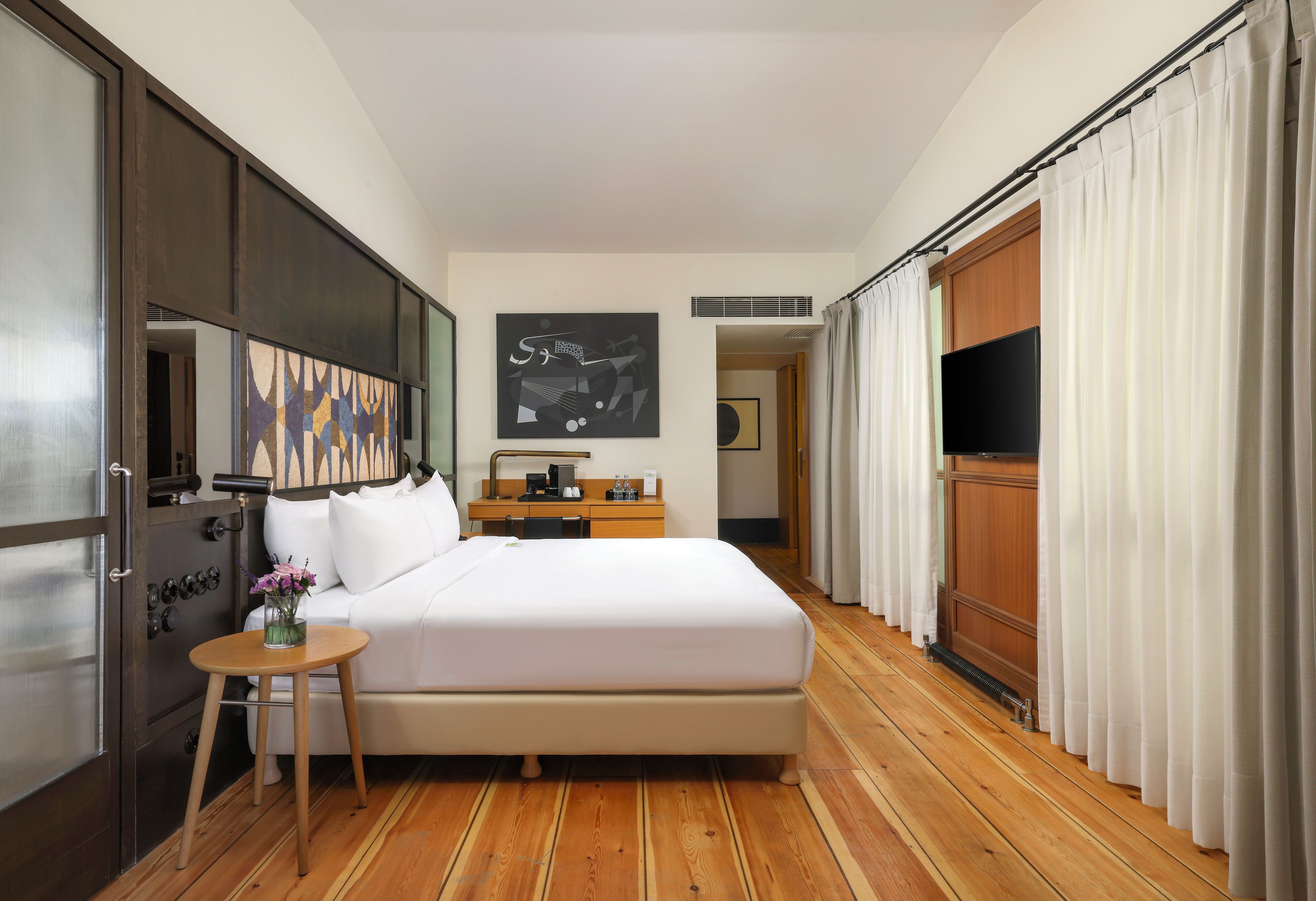 Habitación de hotel con cama, escritorio, silla, pequeña mesa con un jarrón de flores, un cuadro en la pared y televisor.