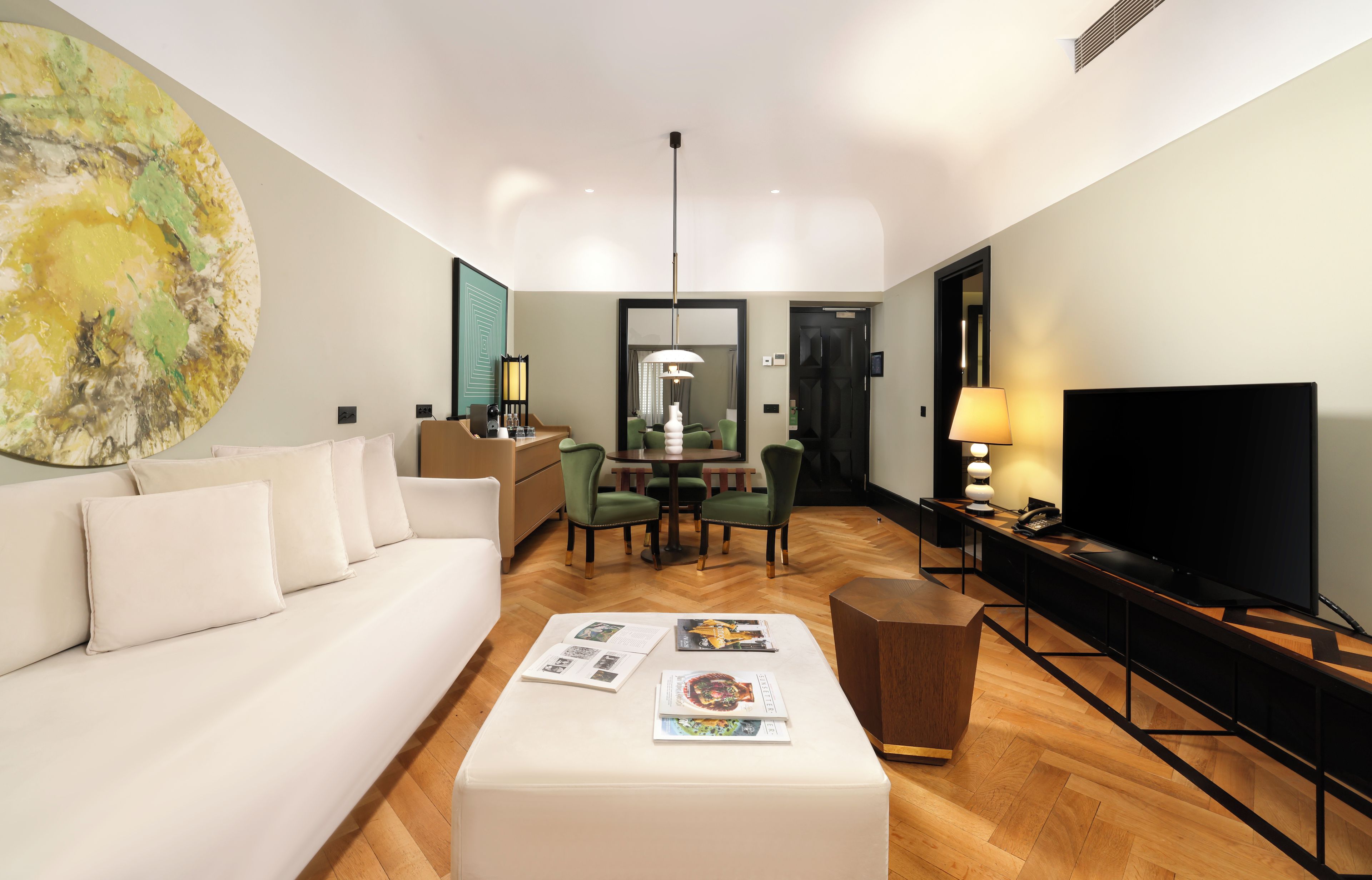 Una habitación de hotel moderna con un sofá blanco, una zona de comedor con sillas verdes, un televisor de pantalla plana y un cuadro abstracto grande en la pared.