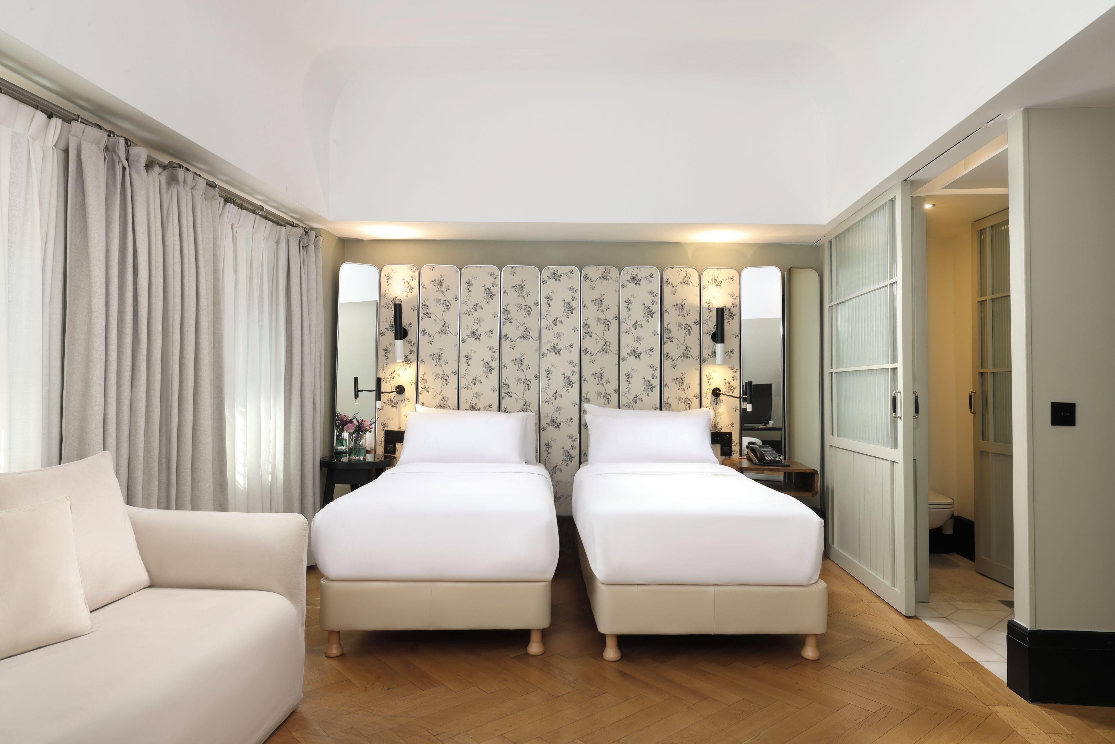 A hotel room with two single beds, a sofa, a nightstand with a lamp, and a mirror.