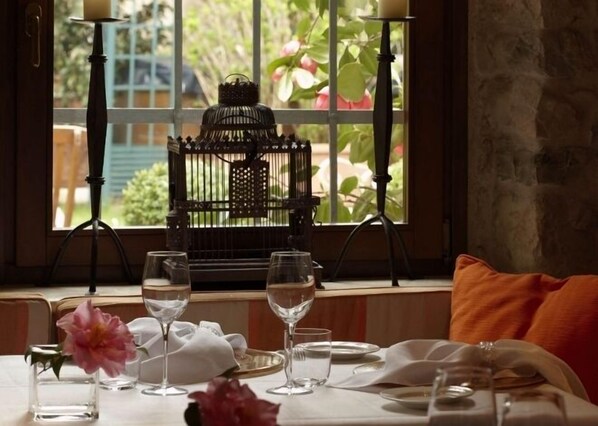 Une table à manger dressée avec des verres à vin et une bougie, près d’une fenêtre donnant sur un paysage verdoyant et une cage à oiseaux.