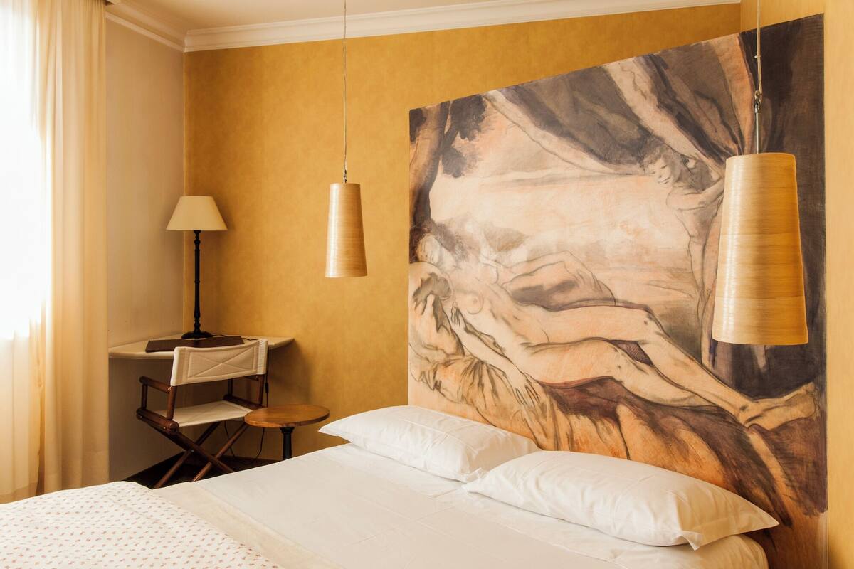 Habitación de hotel con una cama grande, un escritorio con una silla, una lámpara colgante y un cuadro grande en la pared.