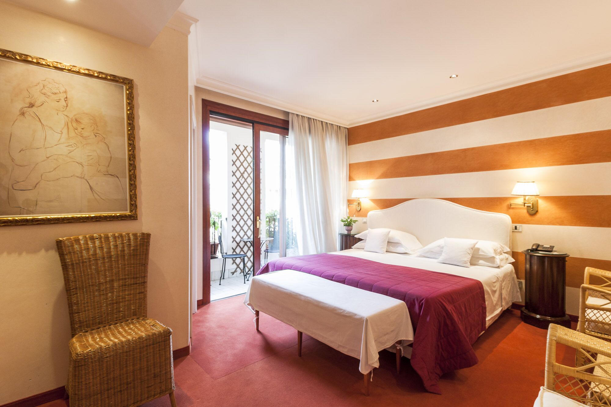 A hotel room with a bed, a bench, a wicker chair, a painting, and a view of the outside.