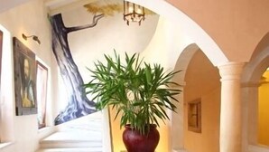 A hallway with a staircase, a large potted plant, and a mural on the wall. - Hotel Dei Chiostri (Follina)