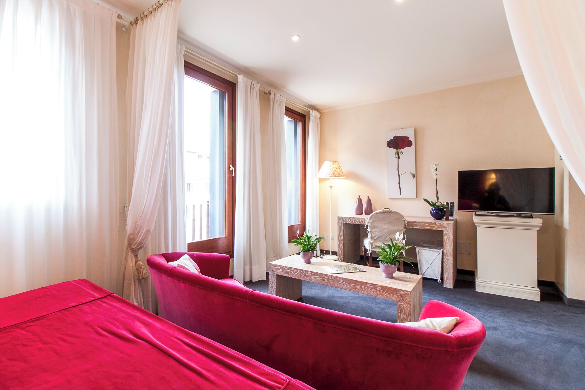 A hotel room with a bed, a desk with a lamp, a TV, and a potted plant.