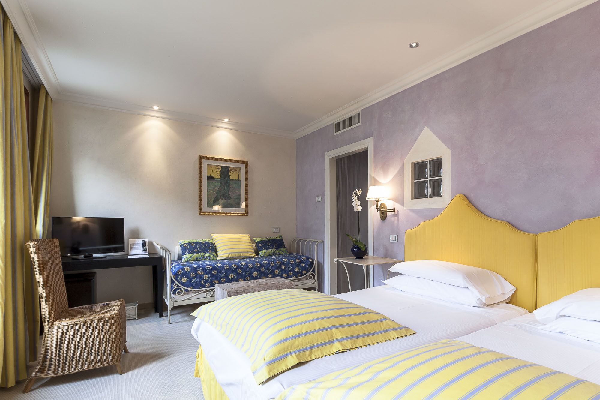 A hotel room with two beds, a desk with a TV, a wicker chair, and a painting on the wall.