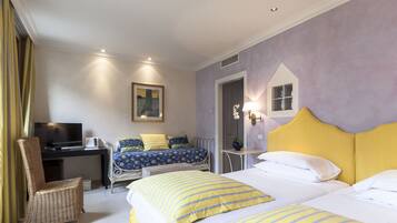 A hotel room with two beds, a desk with a TV, a wicker chair, and a painting on the wall.