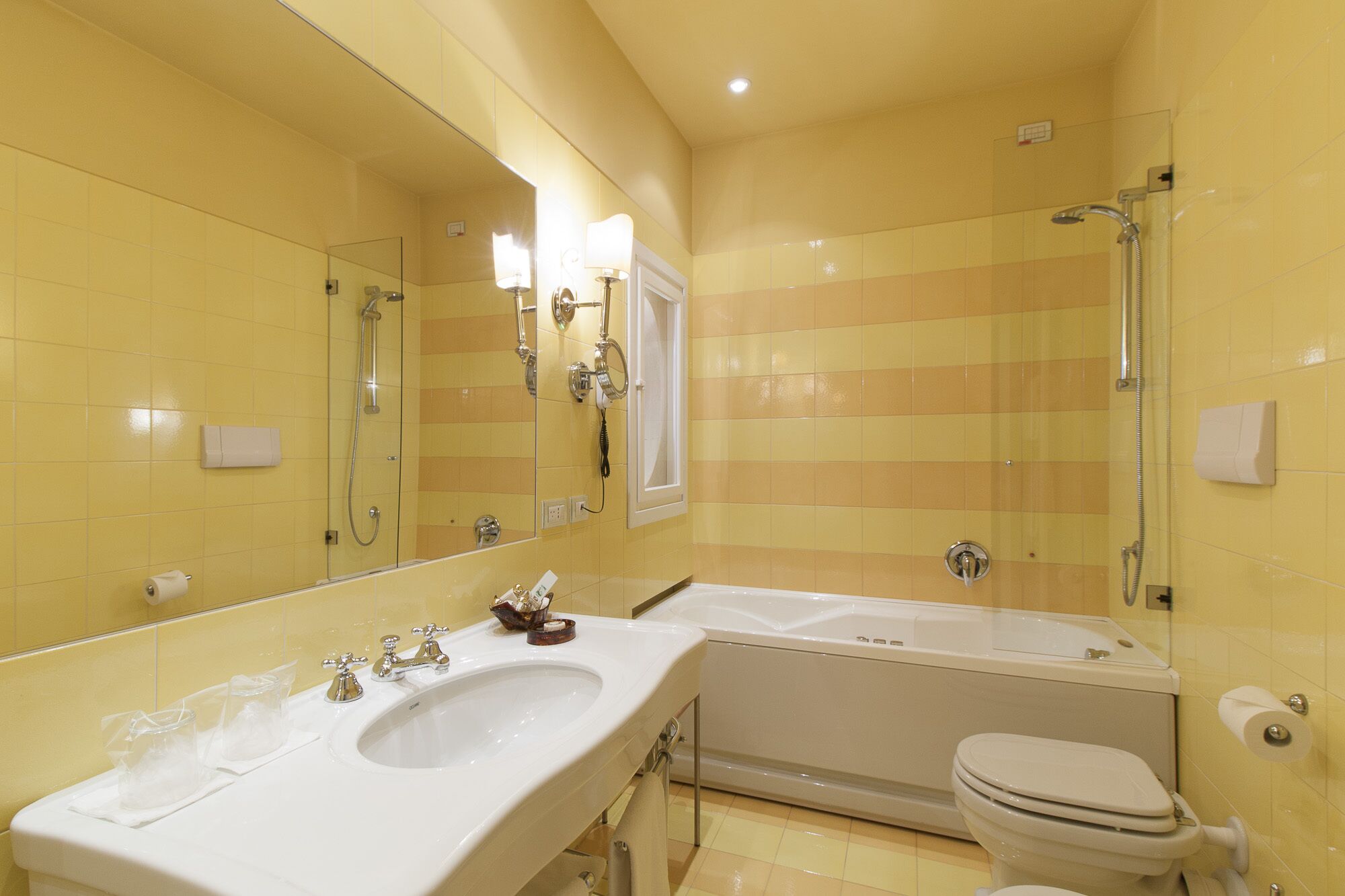 A bathroom with yellow tiled walls, a large mirror, a sink with two basins, a bathtub, and a toilet.