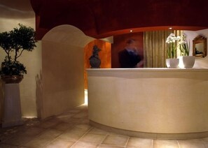 A reception area with a curved reception desk, a potted plant, and a vase with flowers. - Hotel Dei Chiostri (Follina)