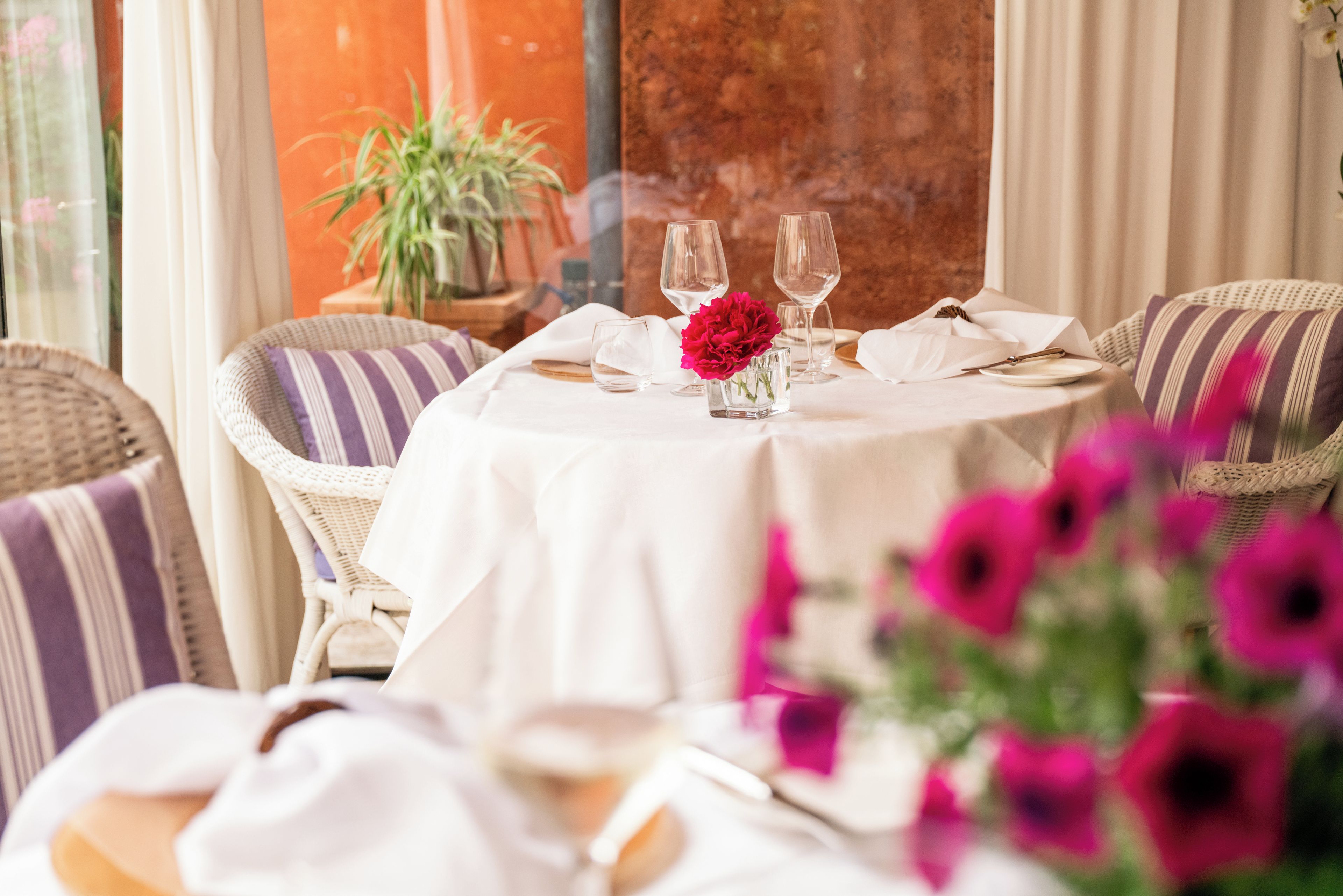 A round table with a white tablecloth, two wine glasses, a small vase with a red flower, and striped cushions on wicker chairs.