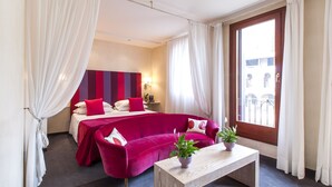 A hotel room with a bed, a red sofa, a wooden coffee table, and a view of the outside. - Hotel Dei Chiostri (Follina)