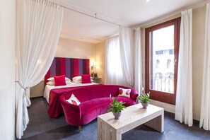 A hotel room with a bed, a red sofa, a wooden coffee table, and a view of the outside.