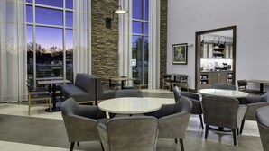 A modern indoor seating area with round tables and wicker chairs, large windows with a view of the outside, and a kitchen area visible through a glass partition.