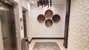 A modern elevator with a metallic finish, a wall-mounted trash bin, and a patterned rug leading to a reception area with a unique textured wall and decorative wall art.