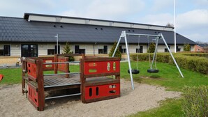 A playground with a swing set and a climbing structure in front of a building with a dark roof. - Danhostel Thorhallen Jels (Rodding)