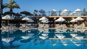 Un área junto a la piscina con sillas de descanso y sombrillas, una palmera y un cielo azul despejado.