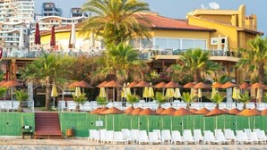 Un hotel frente a la playa con hileras de sombrillas y sillas de descanso, palmeras y un edificio con tejado de tejas rojas.