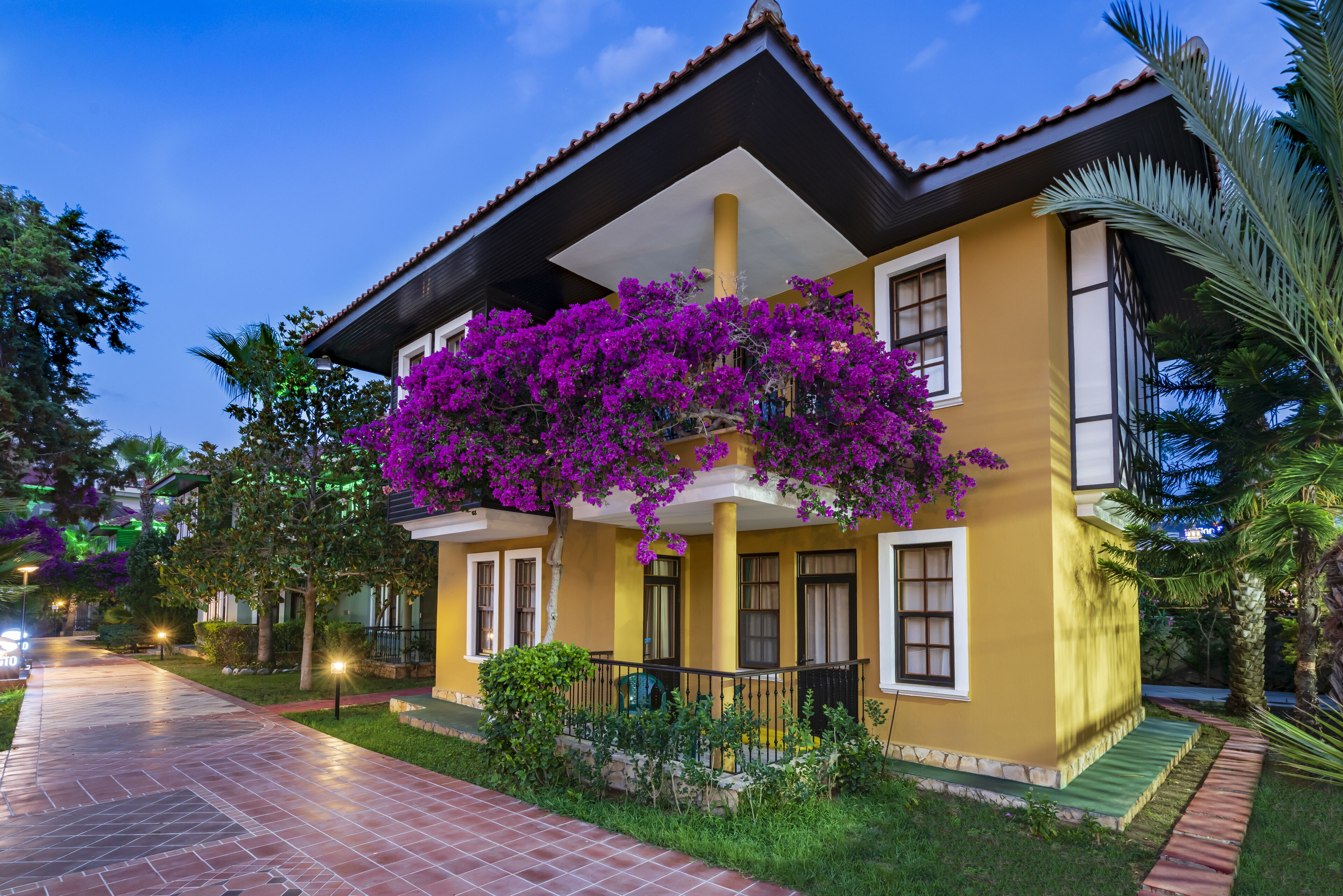 Um edifício de dois andares com fachada amarela, uma varanda com flores roxas e um caminho de azulejos que leva à entrada.