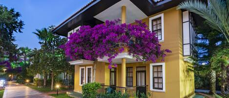 Um edifício de dois andares com fachada amarela, uma varanda com flores roxas e um caminho de azulejos que leva à entrada.