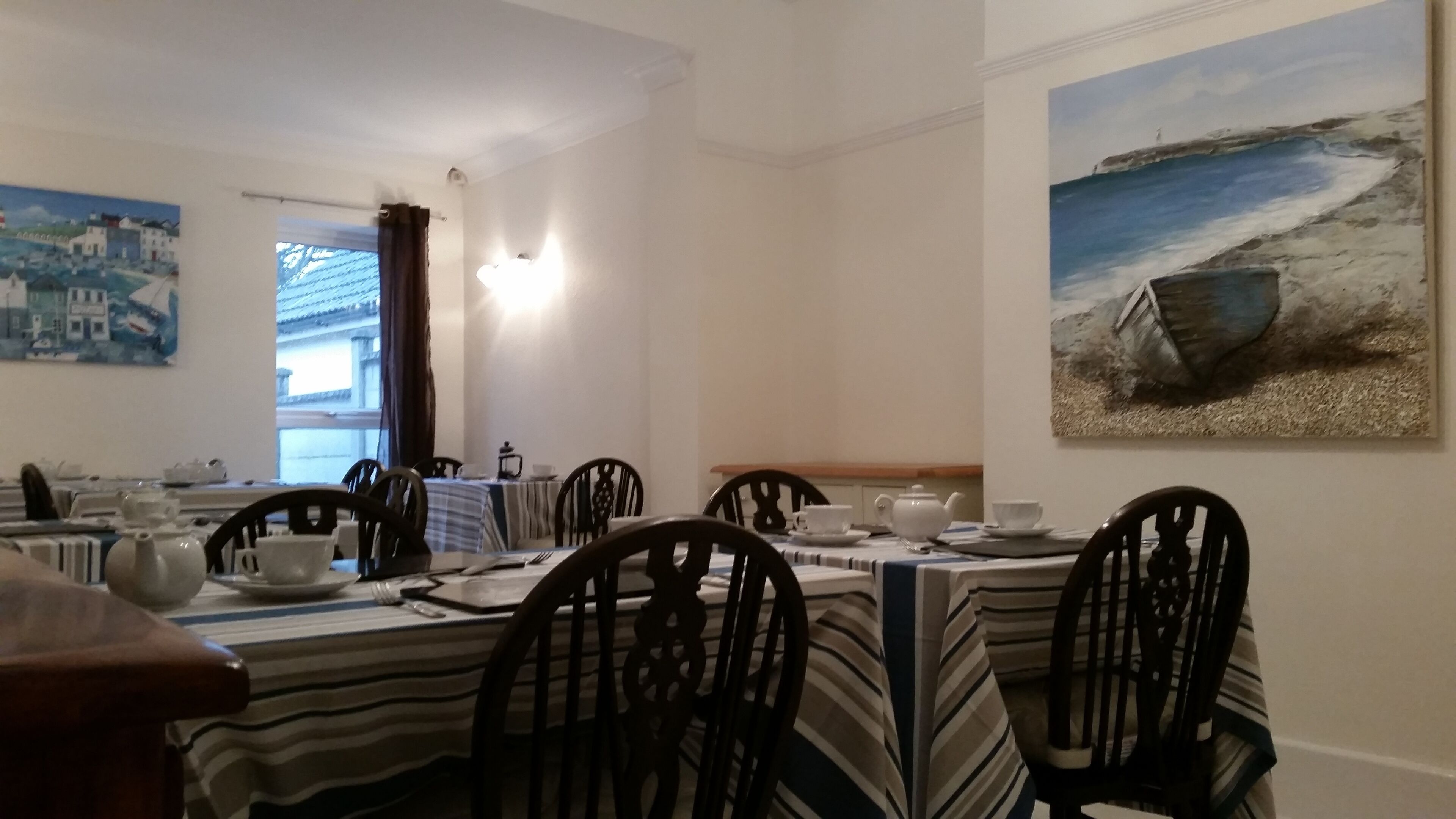 A dining area with striped tablecloths, wooden chairs, and a painting of a seascape.