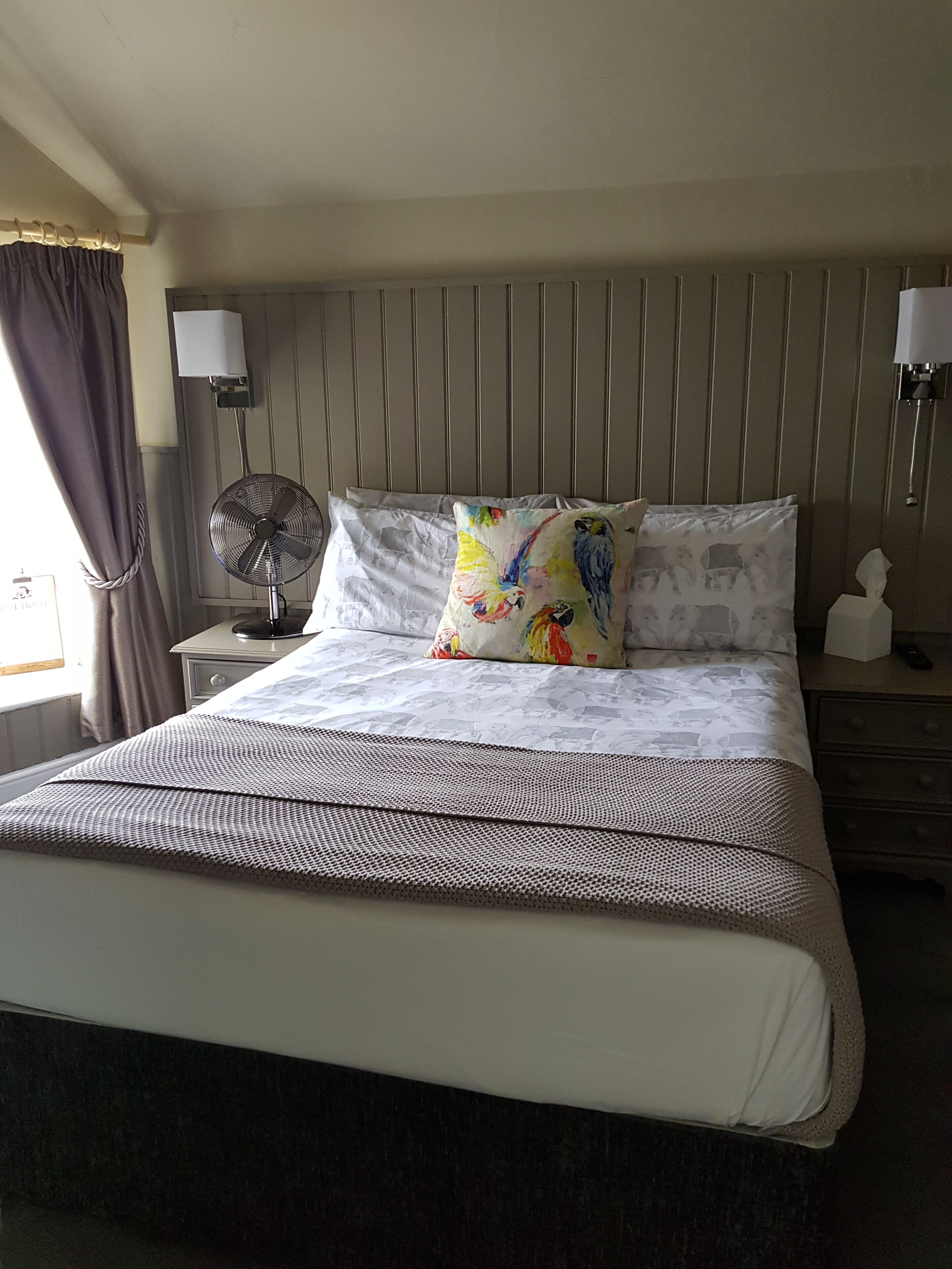 A neatly made bed with patterned bedding, a standing fan, and a bedside table with a lamp.