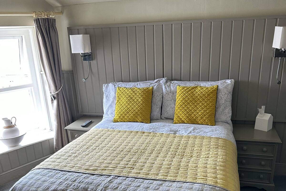 A neatly made bed with a quilted headboard, a bedside table with a lamp, and a window with curtains.