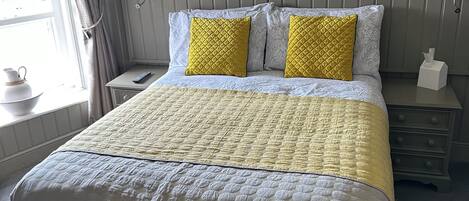 Cama bem arrumada com cabeceira acolchoada, mesa de cabeceira com abajur e janela com cortinas.