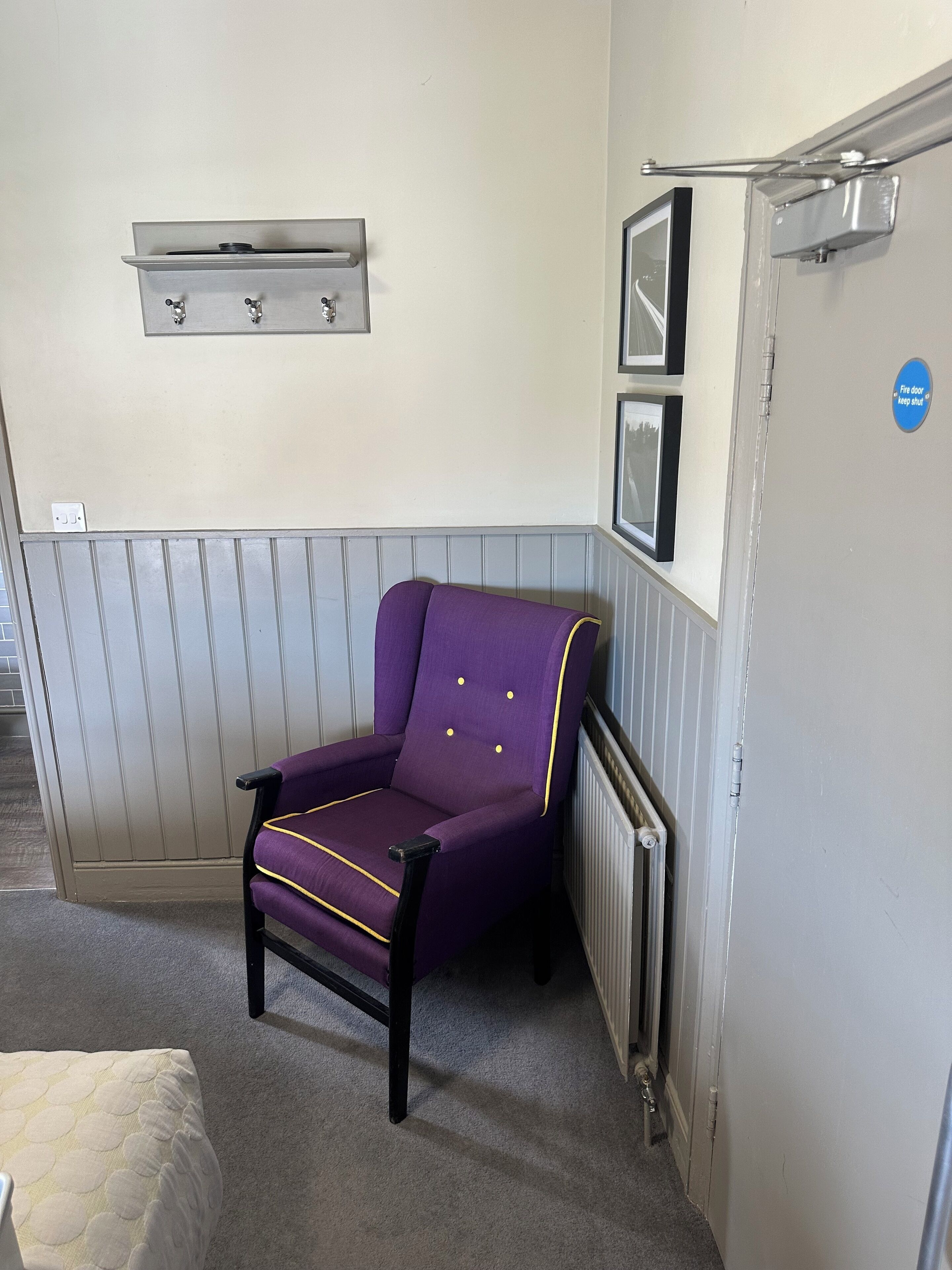 A purple armchair in a corner with a radiator, a fire door sign, and a shelf above it.