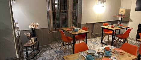 Un comedor con una mesa de madera preparada para cuatro personas, rodeada de sillas naranjas, un piso de baldosas estampadas y una ventana con persianas.