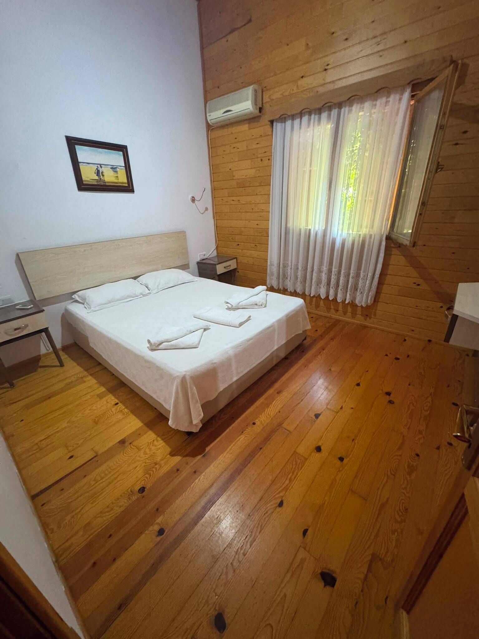 Un dormitorio con piso de madera, una cama con ropa blanca, una mesita de noche, un cuadro en la pared y una ventana con cortinas.