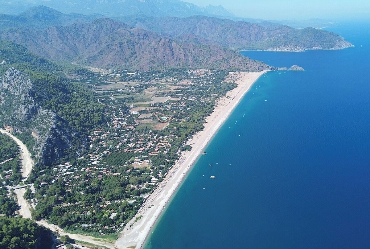 An aerial view of a coastal town with a long beach, dense greenery, and mountainous terrain in the background.