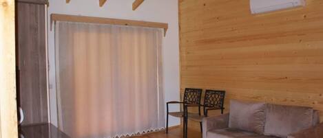 Una habitación con paneles de madera, un sofá, dos sillones, una ventana con cortinas y techo de madera.