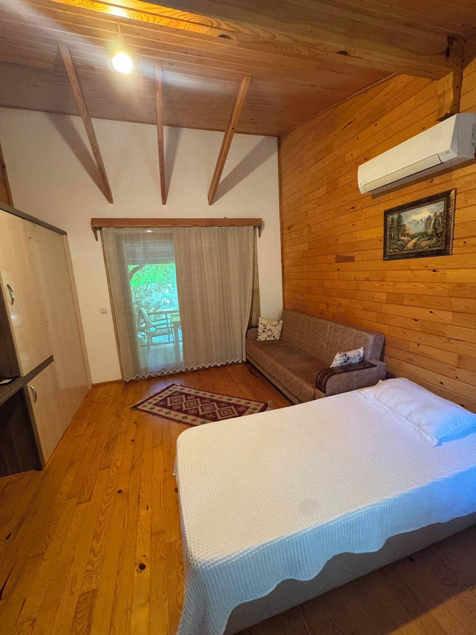 A room with a bed, a sofa, a wooden floor, a window with curtains, and an air conditioning unit.
