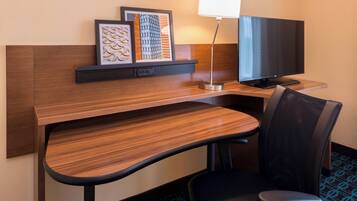 A hotel room with a wooden desk, a black office chair, a flat-screen TV, a desk lamp, and framed artwork on the wall.