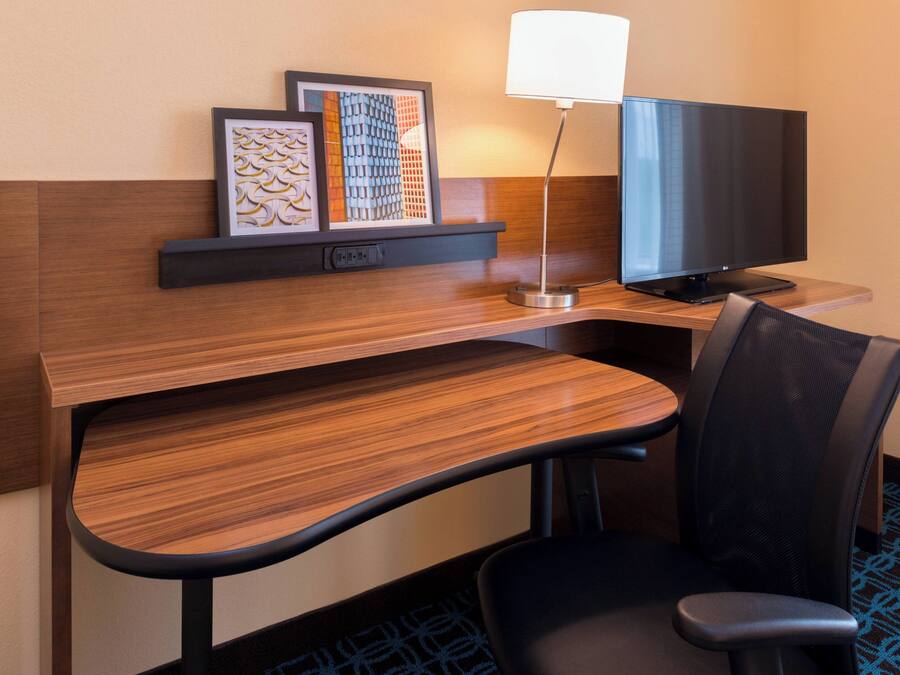 A hotel room with a wooden desk, a black office chair, a flat-screen TV, a desk lamp, and framed artwork on the wall.