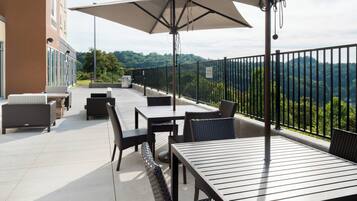 An outdoor patio with tables and chairs, a large umbrella, and a view of a forested area.