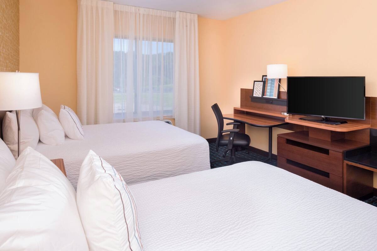 Ein Hotelzimmer mit zwei Betten, einem Schreibtisch mit Fernseher und einem Fenster mit Vorhängen.