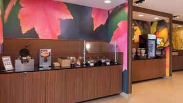 A hotel reception area with a wooden counter, a waffle maker, a coffee machine, and a fruit display.