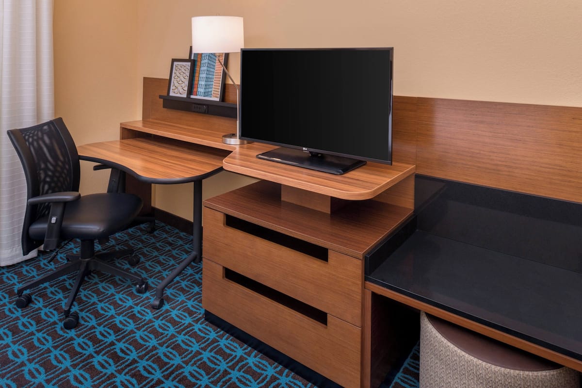 Ein Hotelzimmer mit Schreibtisch aus Holz, einem schwarzen Bürostuhl, einem Flachbildfernseher, einer Lampe und einem gerahmten Bild auf dem Schreibtisch.