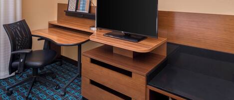 A hotel room with a wooden desk, a black office chair, a flat-screen TV, a lamp, and a framed picture on the desk.