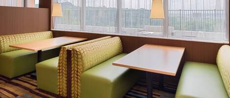 A hotel room with a seating area featuring green cushioned benches and a table, large windows with sheer curtains, and a striped carpet.