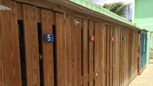 Een rij houten locker met genummerde deuren, een groen gebouw op de achtergrond en een geplaveid pad.