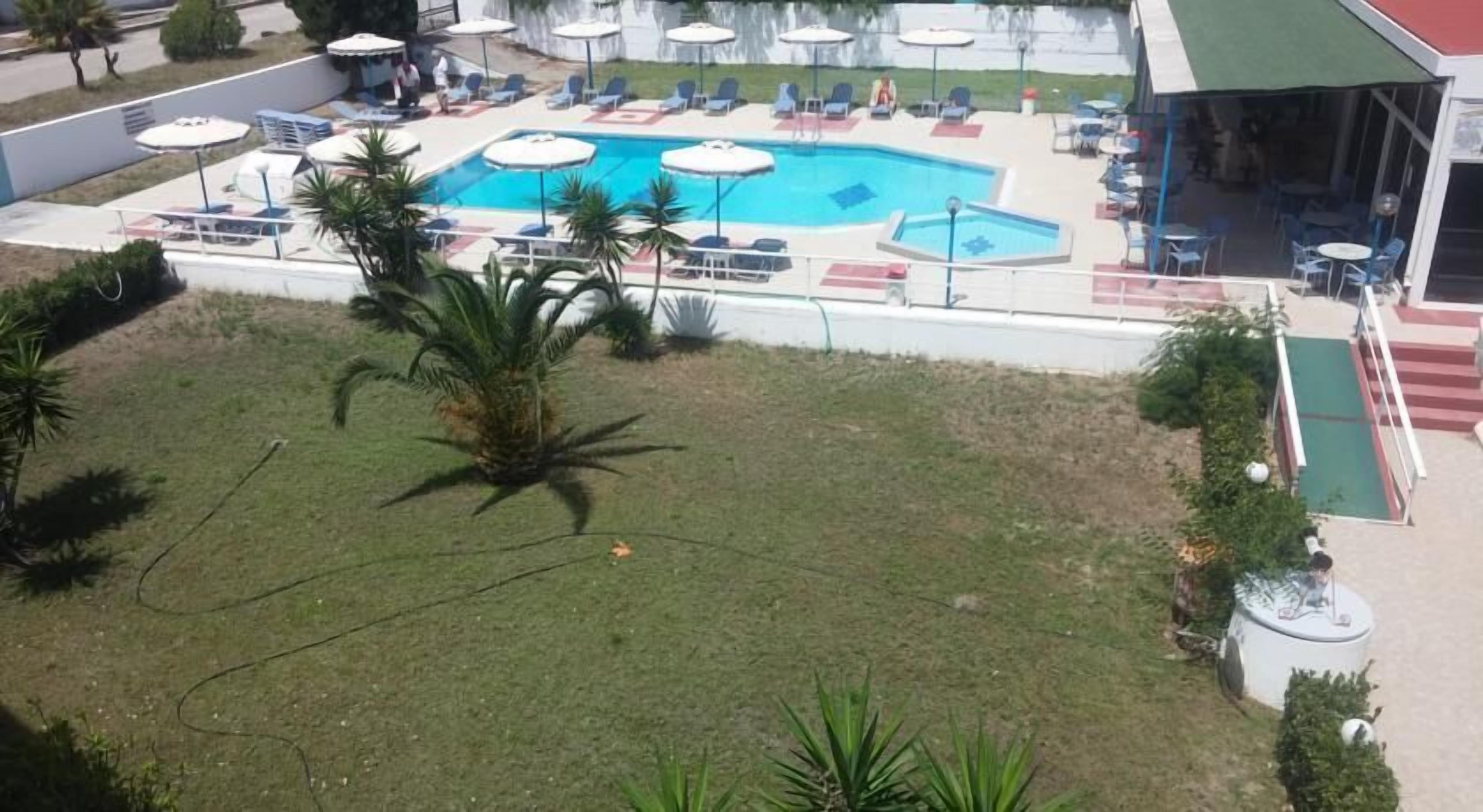A pool area with lounge chairs and umbrellas, surrounded by a grassy area with palm trees.
