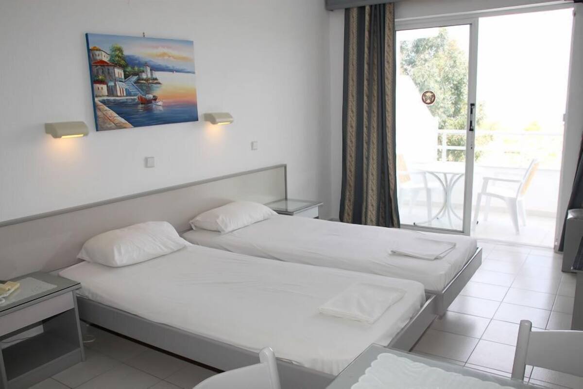A hotel room with two single beds, a painting of a coastal scene, a sliding glass door to an outdoor area with a table and chairs, and a small desk with a lamp.