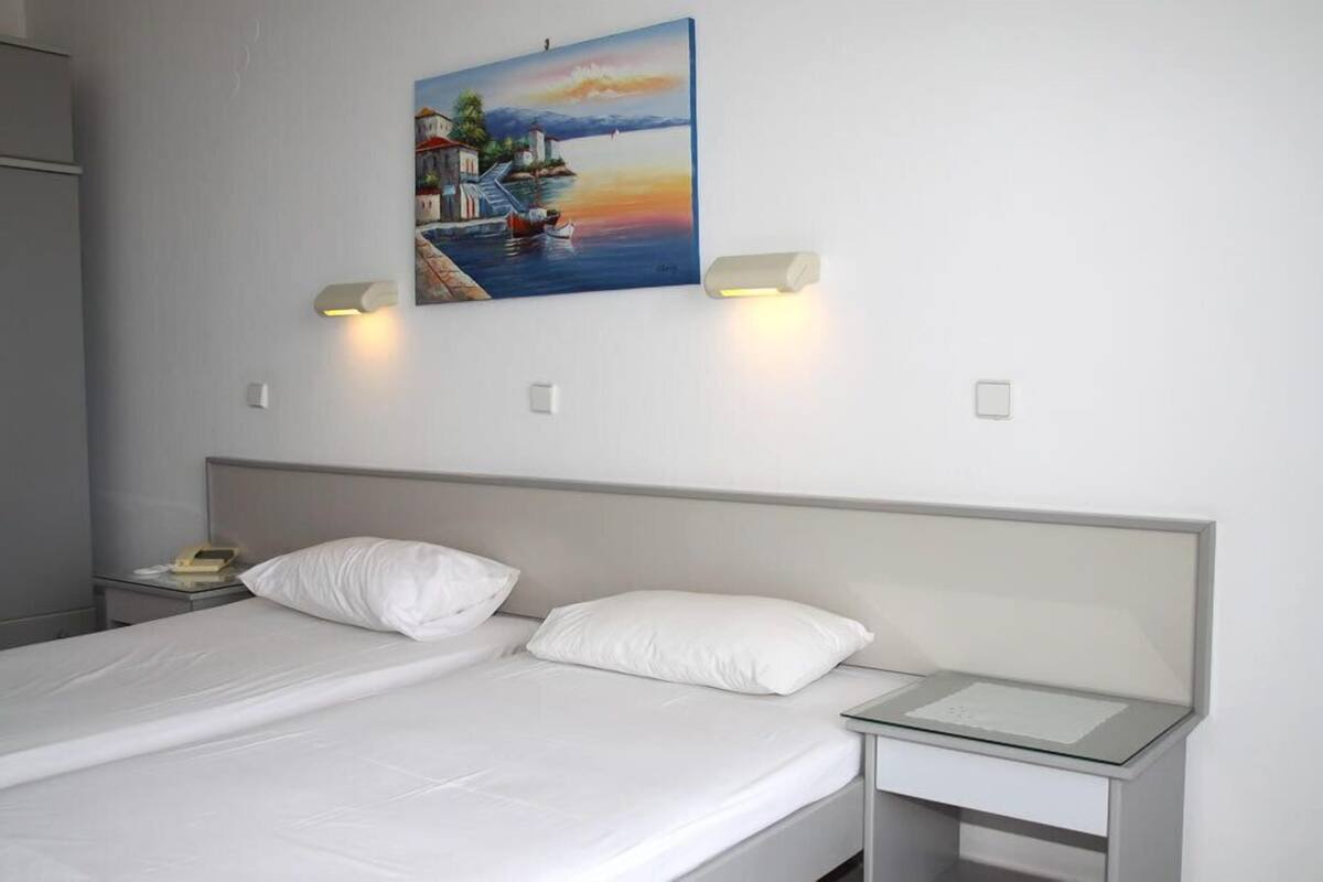 A neatly made bed with white linens, a bedside table with a lamp, and a painting of a coastal scene on the wall.