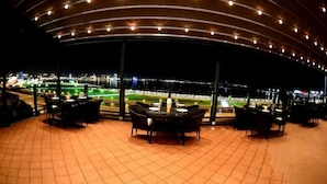 An outdoor dining area with a curved roof, tables, and chairs, overlooking a lit-up sports field. - Riviera Hotel  (Baku)