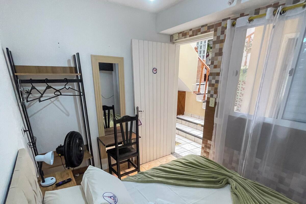 A hotel room with a bed, a chair, a desk, a fan, and a window with curtains.