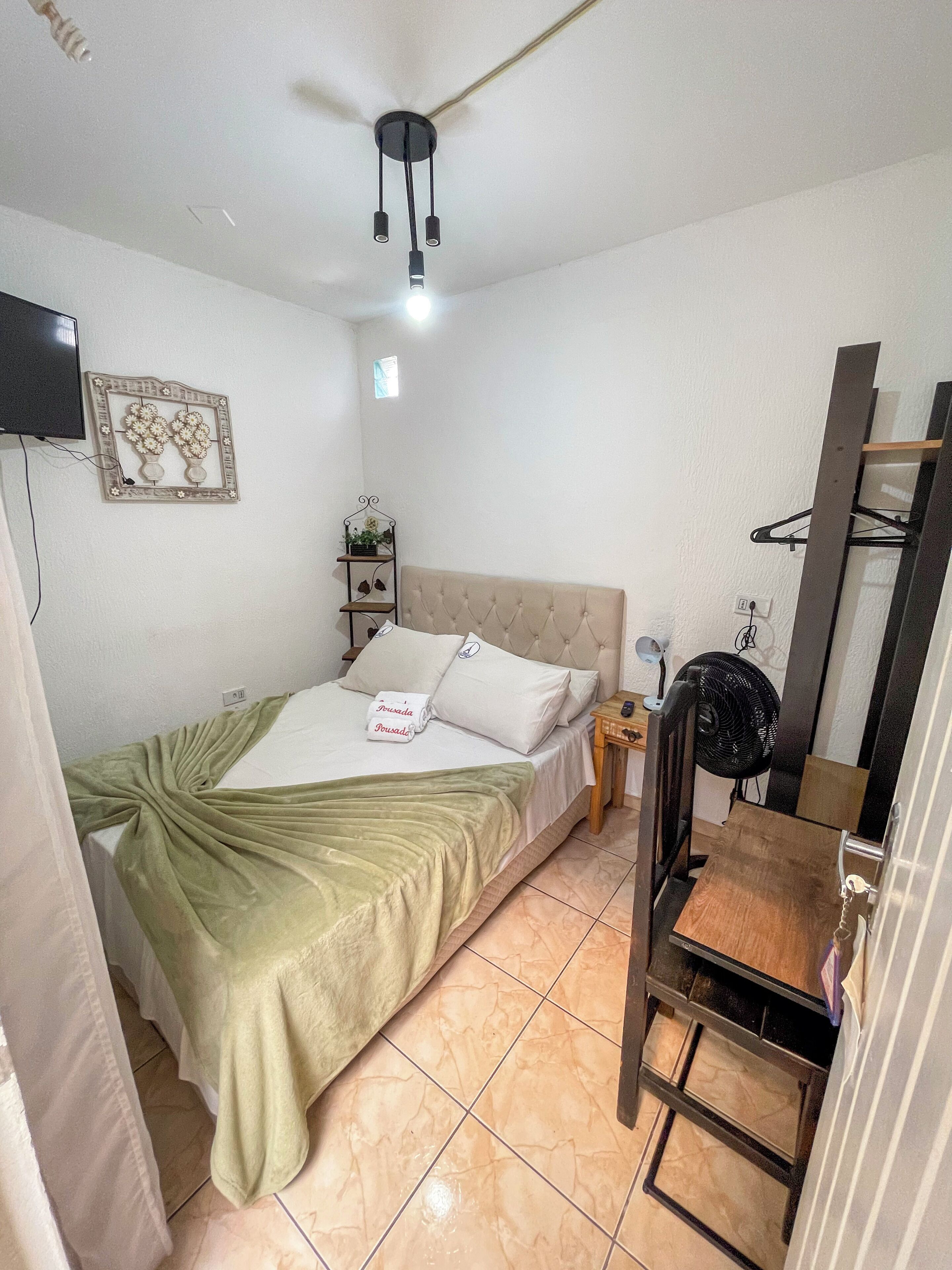 A bedroom with a bed, a bedside table, a fan, a TV, and a wall-mounted shelf.