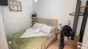 A bedroom with a bed, a bedside table, a fan, a TV, and a wall-mounted shelf.